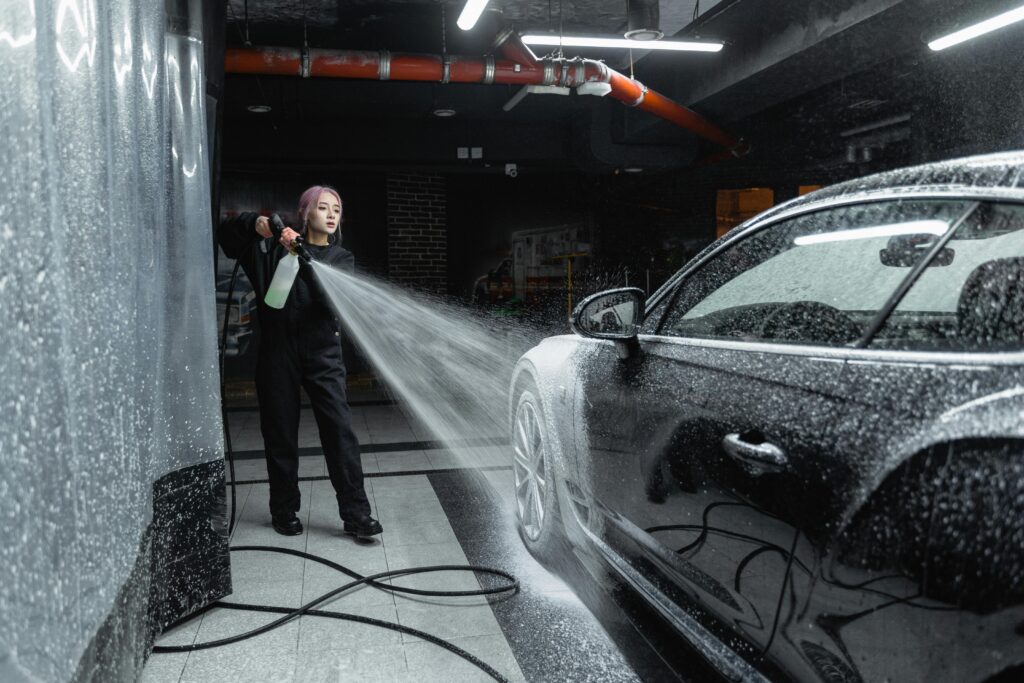 Green Car Wash: Eco-Friendly Mobile Detailing at Your Doorstep Woman using a pressure washer to clean a black car in an indoor car wash setting.