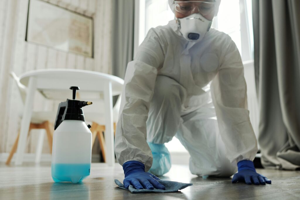 Person in protective gear cleaning a floor with disinfectant spray and cloth indoors.