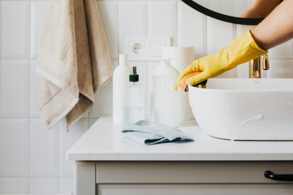 Bring Sparkle and Freshness to Every Corner Person cleaning a bathroom sink with yellow gloves and cleaning products on a countertop.