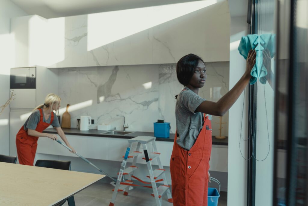 Kitchen Deep Cleaning Two women cleaning a modern kitchen, wiping glass surfaces and mopping the floor.